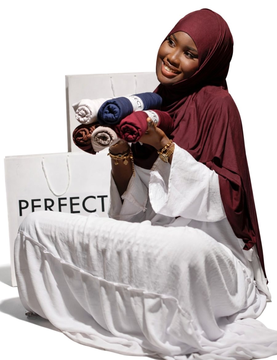 Femme souriante en hijab bordeaux, assise devant des sacs Perfect Hijab, tenant des hijabs roulés